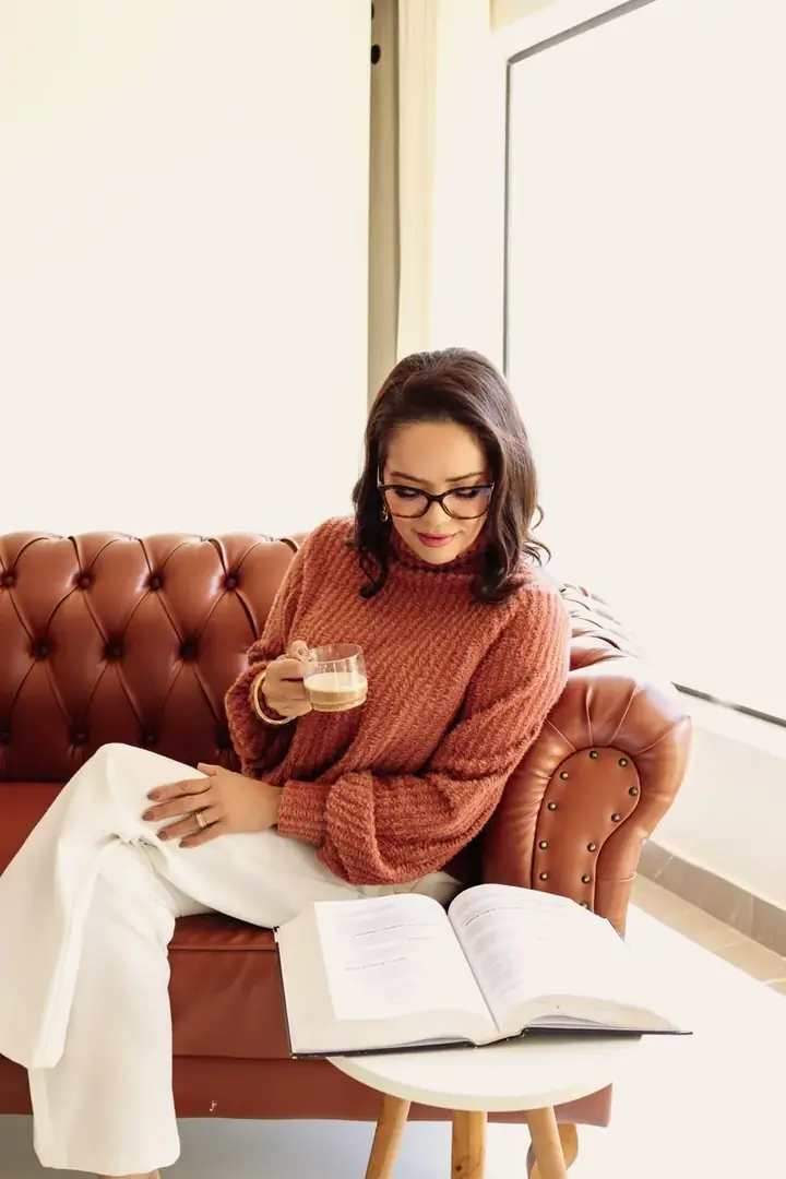 Cristia Melisa sentada em um sofá de couro, lendo um livro com uma xícara de café